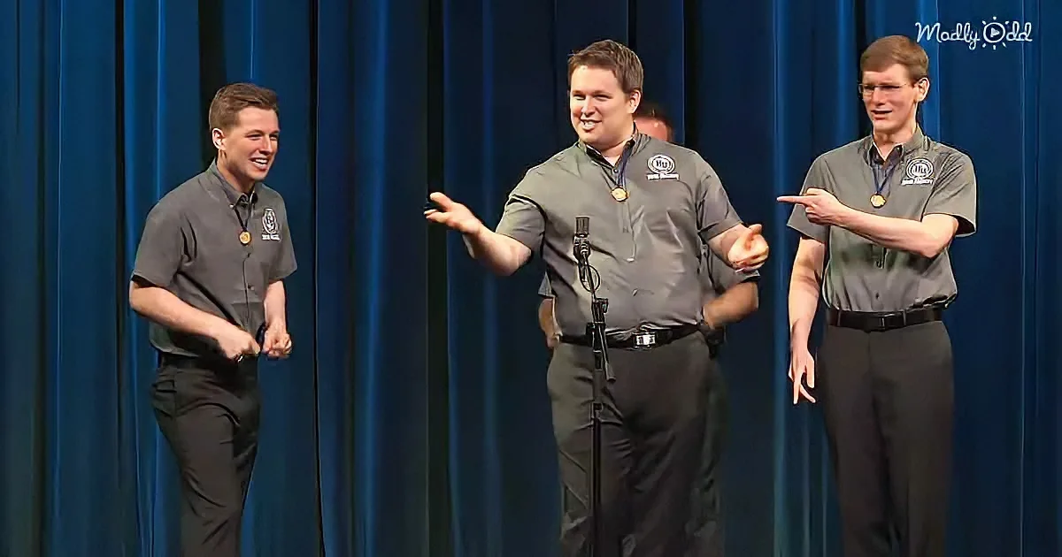 Three men in uniform stand on stage, one gesturing with hands, another laughing, third pointing, all wearing medals.