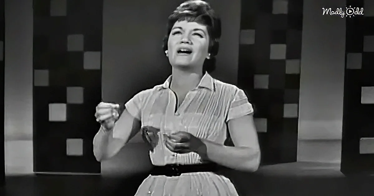 A woman in a 1950s dress holds a glass, singing with a microphone, against a checkered backdrop.