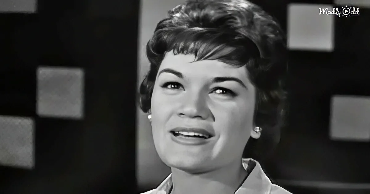 A woman with short, styled hair and pearl earrings smiles softly, wearing a light-colored shirt.