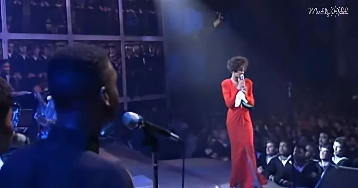 A woman in a red dress sings on stage, surrounded by an audience and a spotlight.