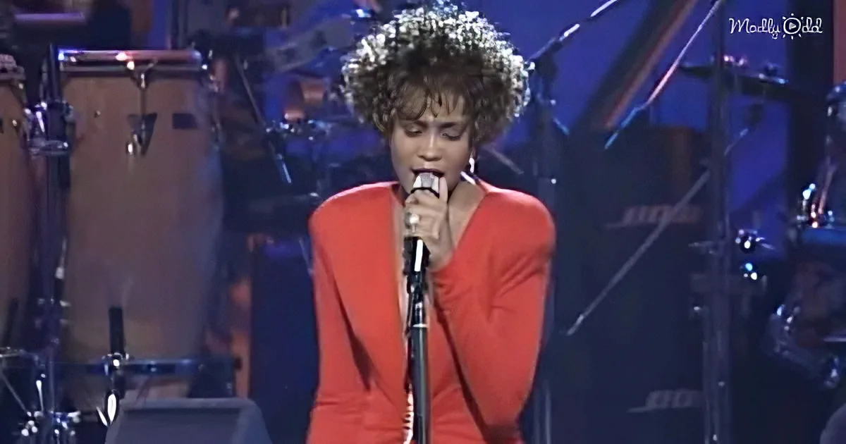 A woman in a red outfit sings into a microphone on stage with a band in the background.