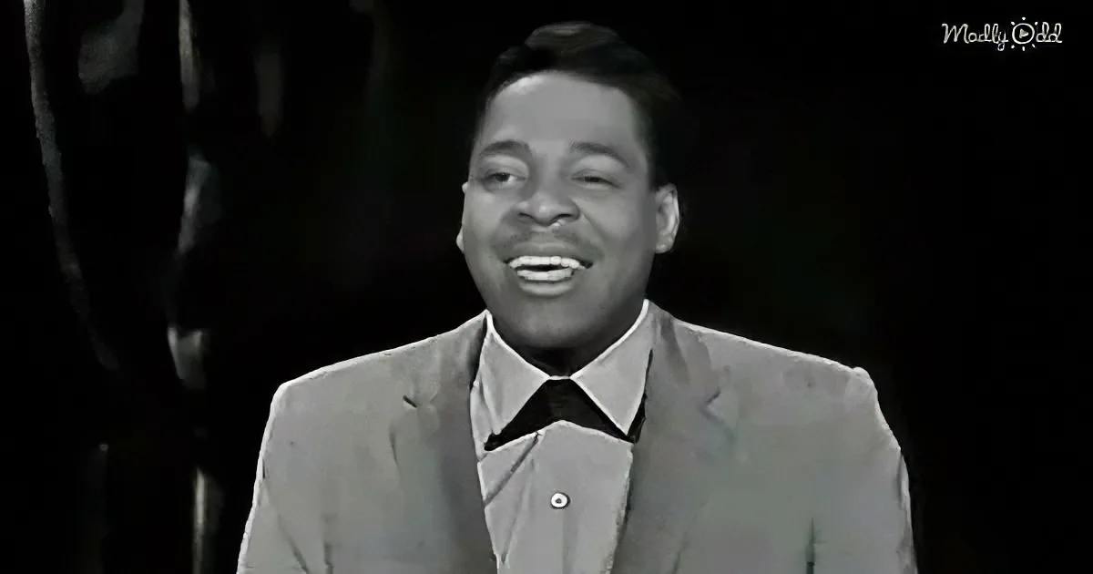 A man in a suit and tie smiles widely, wearing a black bow tie, against a dark background.