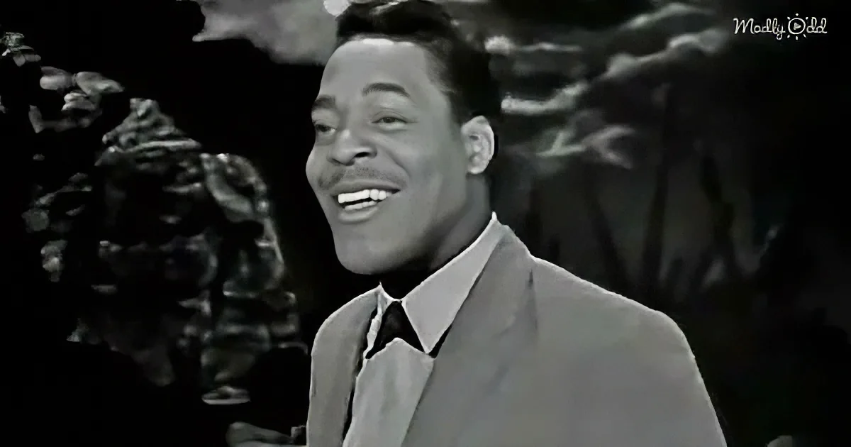 A man in a suit and tie smiles warmly, with a blurred background of foliage.