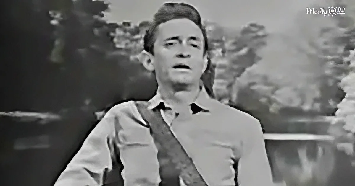 A man in a light-colored shirt and tie stands outdoors, possibly singing or speaking, with a blurred background.