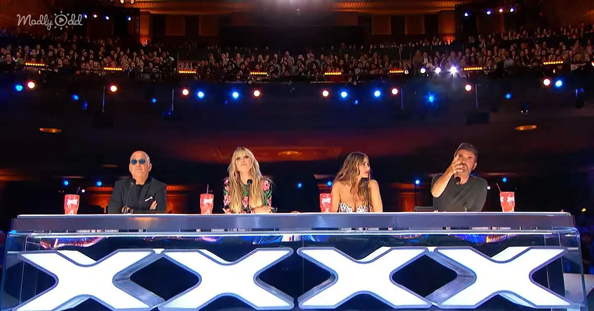 Four judges sit at a table with "XXX" illuminated, in front of a large audience in a dimly lit venue.