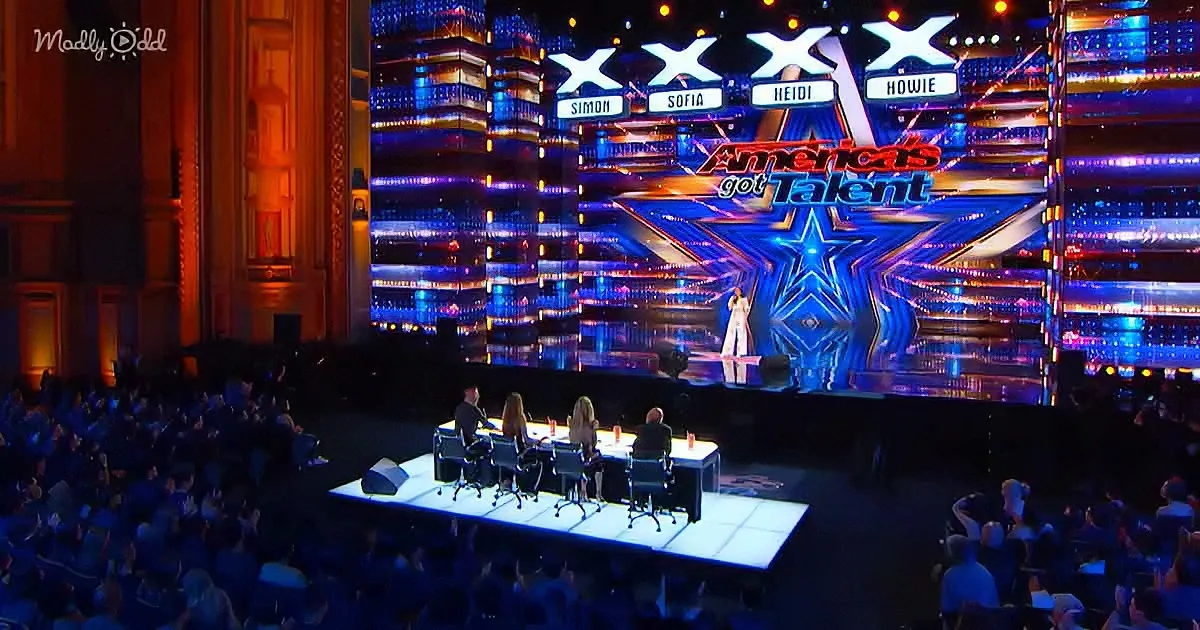 A performer stands on a brightly lit stage with judges seated at a table in front of a large audience.