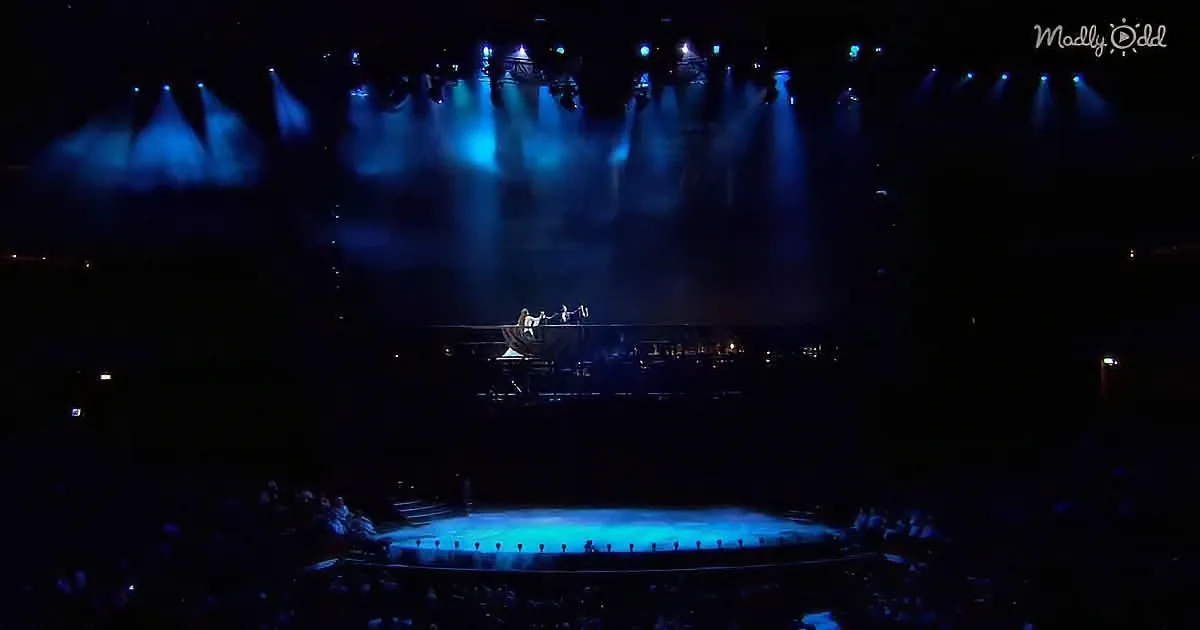 A performer stands on a raised platform under dramatic blue stage lights, surrounded by a darkened audience.