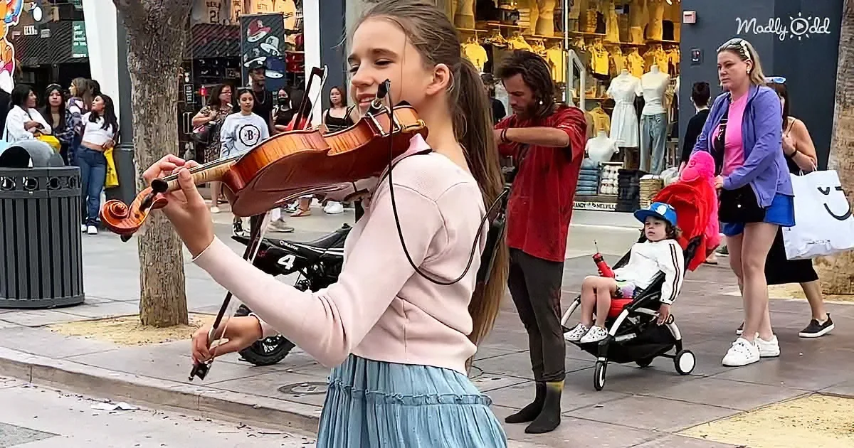 A young girl plays violin on a busy sidewalk, surrounded by pedestrians and a colorful street scene.