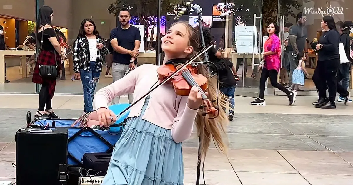 A young woman in a blue skirt plays the violin in a public space, surrounded by onlookers and a store in the background.