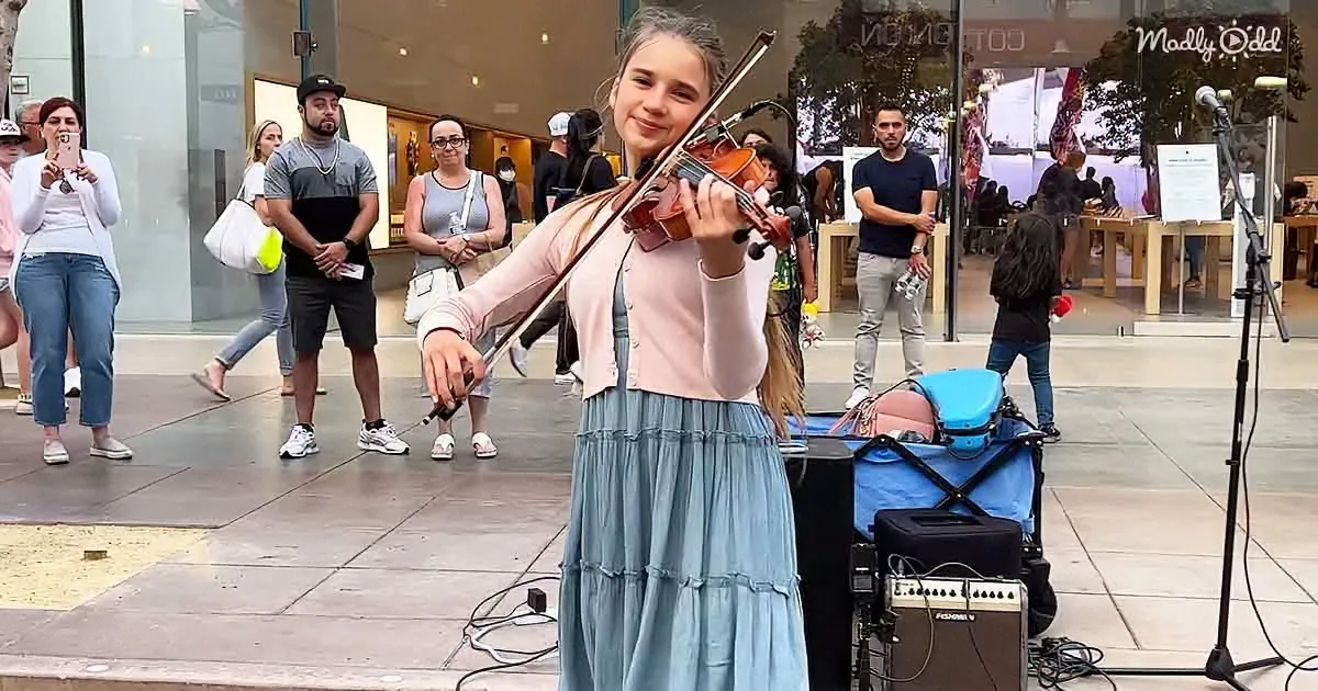 A young woman plays violin on a city street, surrounded by onlookers and musical equipment.