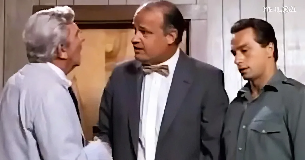 A man in a suit and bow tie is talking to another man in a white shirt, with a third man in a gray shirt observing.