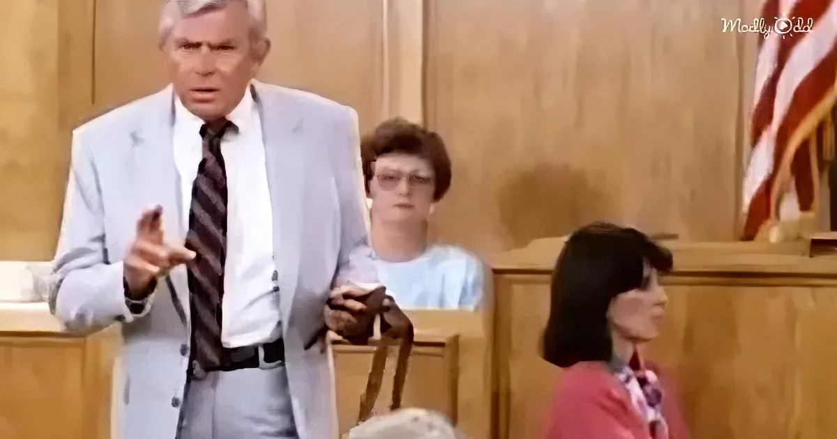 A man in a suit gestures emphatically in a courtroom setting, with a woman in the background and an American flag visible.
