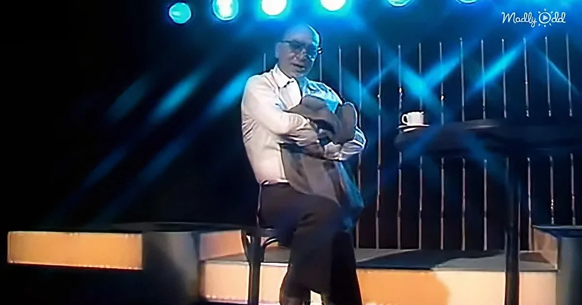 A man sits on a chair on stage, holding a guitar, with blue stage lights behind him.