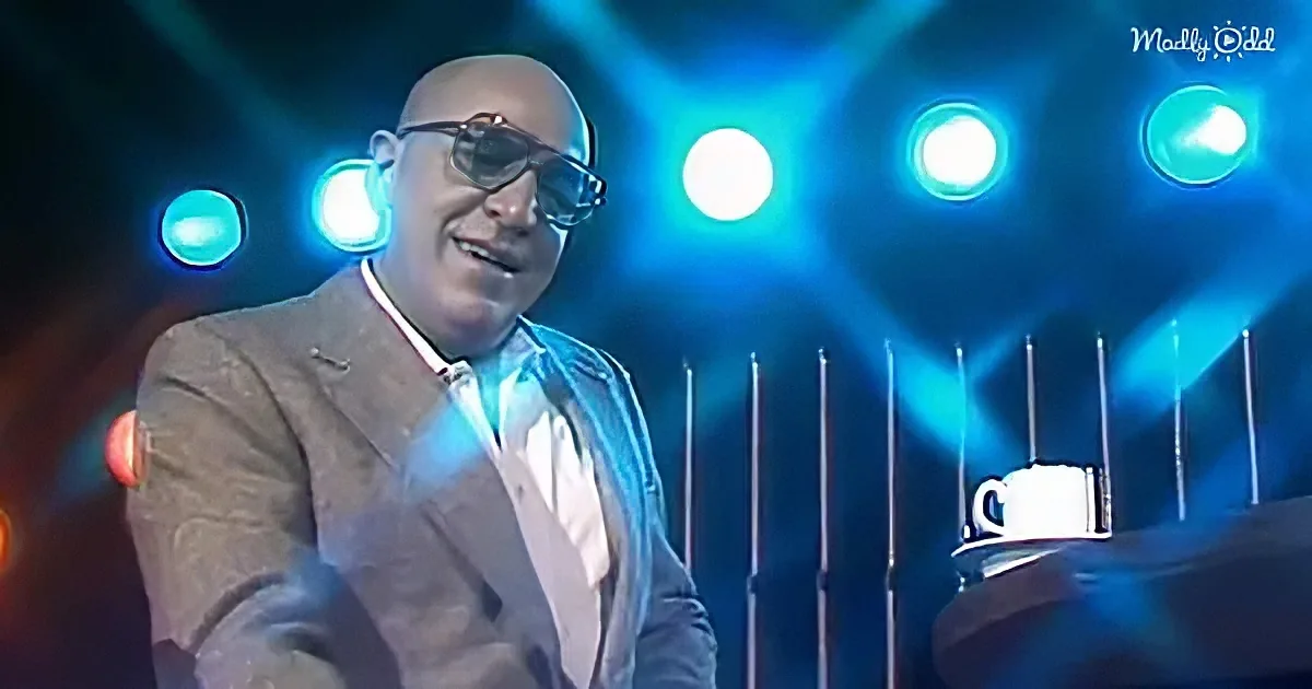 A man in a suit and glasses smiles on stage with blue lights and a mug on a table.
