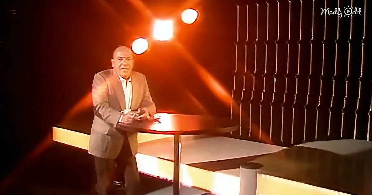 A man in a suit stands at a desk, illuminated by stage lights, with a dark, textured backdrop.