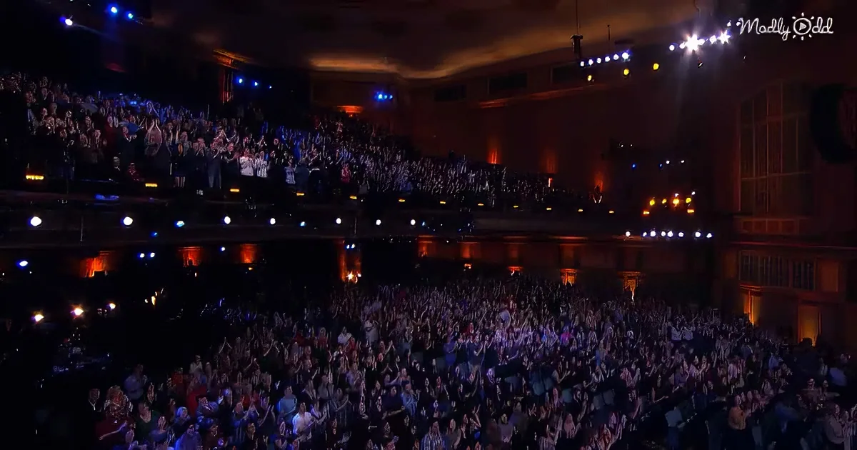A large, enthusiastic audience in a theater claps and cheers under colorful stage lights.