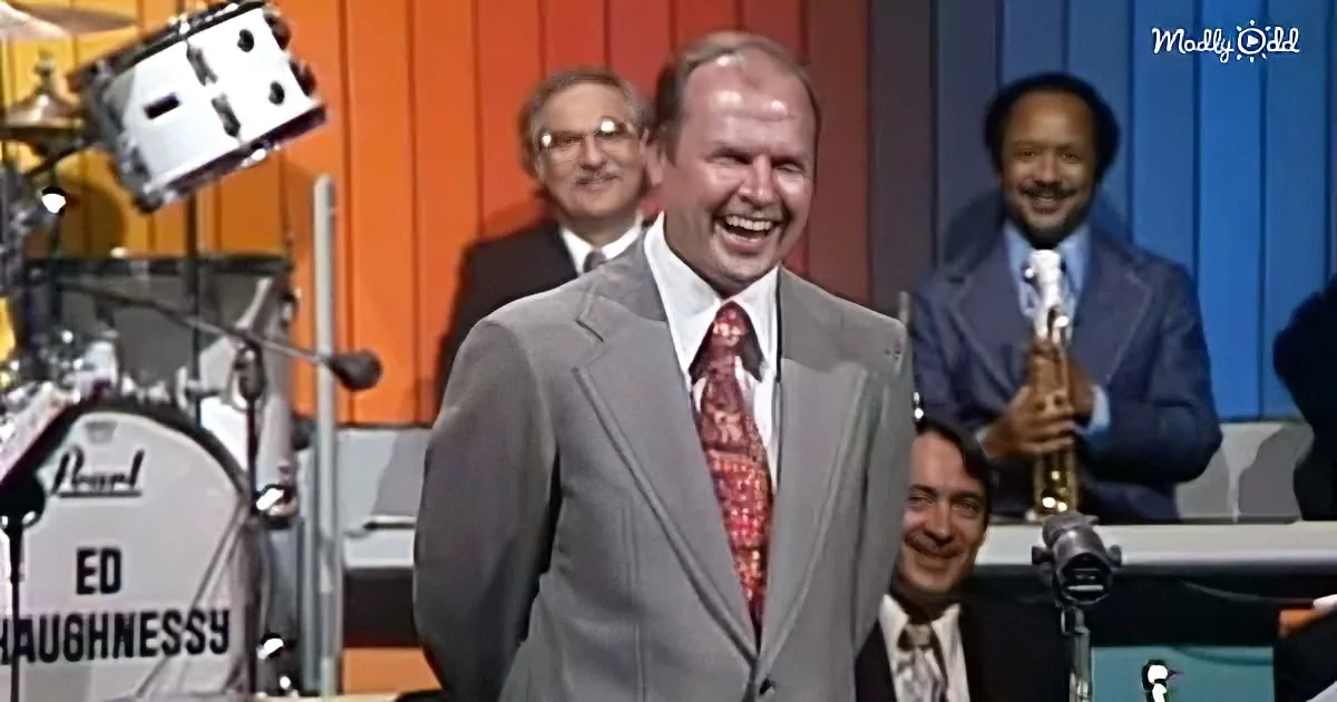 A man in a gray suit and red tie laughs on stage with a band behind him.