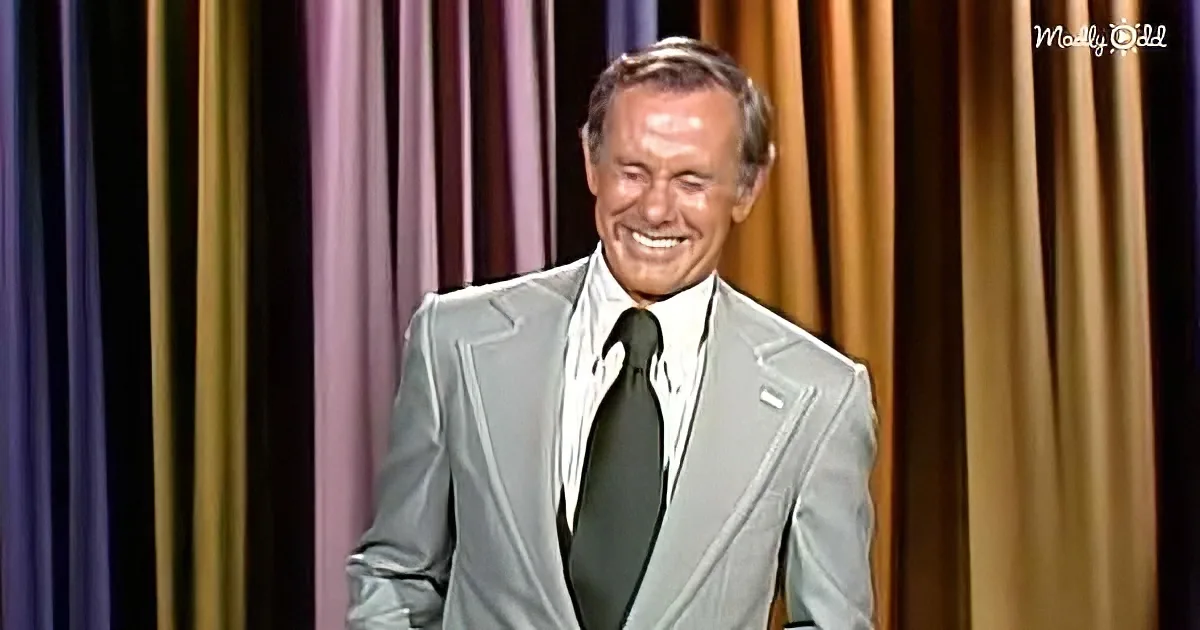 A man in a light gray suit and dark tie smiles on stage with purple and gold curtains behind him.