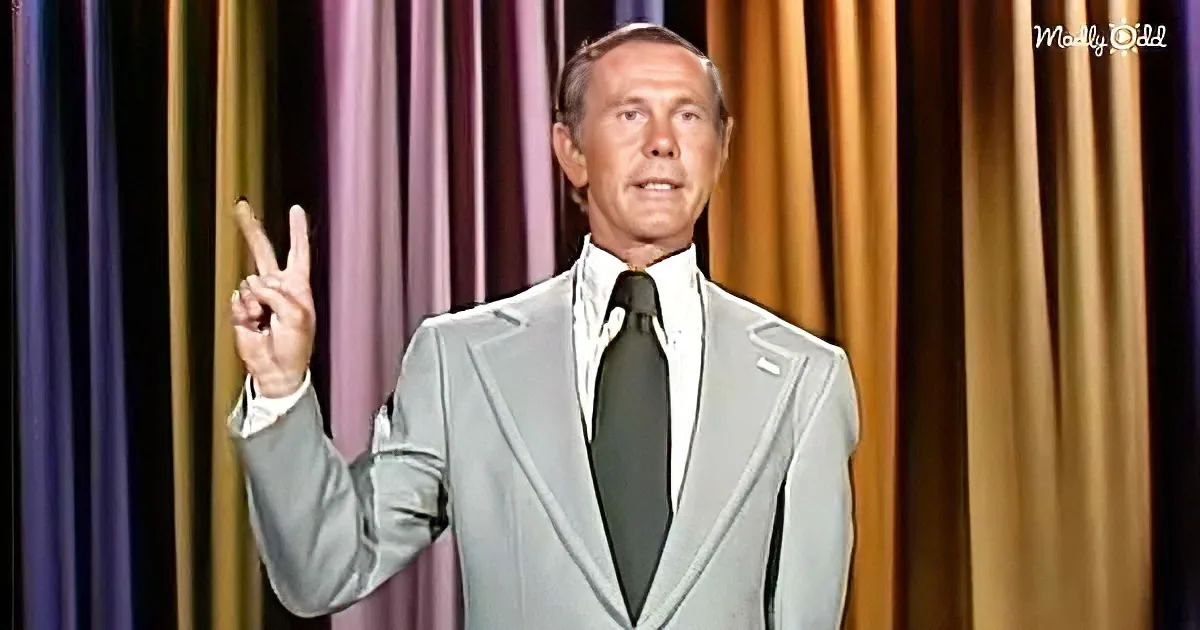 A man in a light gray suit and white shirt makes a peace sign against a colorful curtain backdrop.