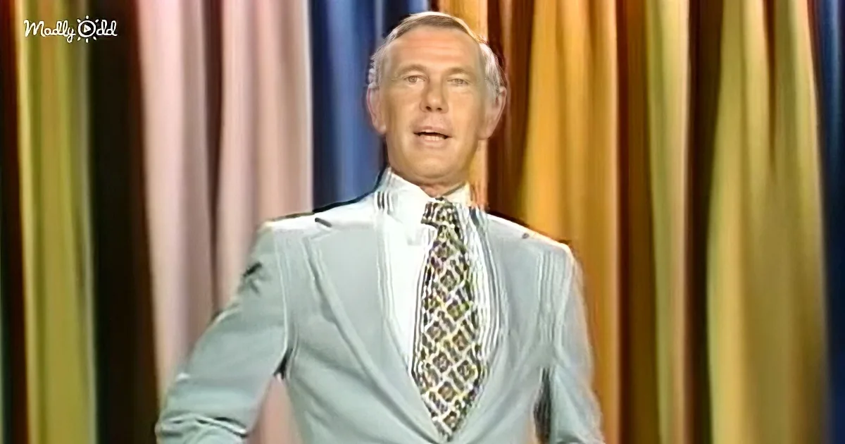 A man in a light blue suit and patterned tie stands against a colorful curtain backdrop.