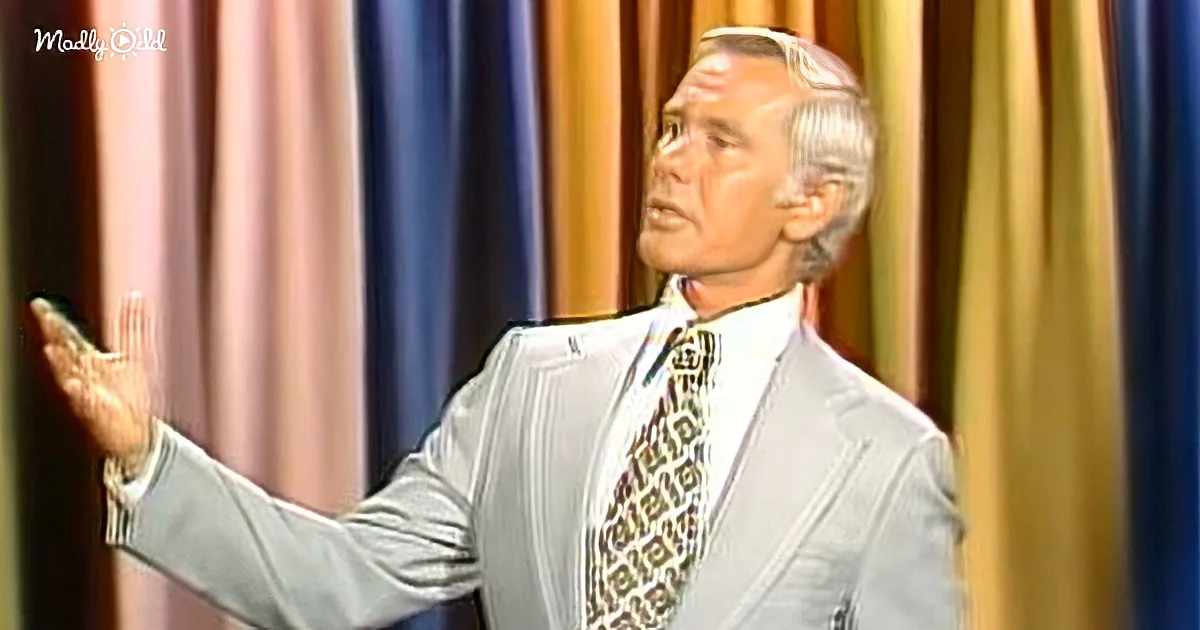 An older man in a light gray suit and patterned tie gestures with his right hand, standing in front of colorful curtains.