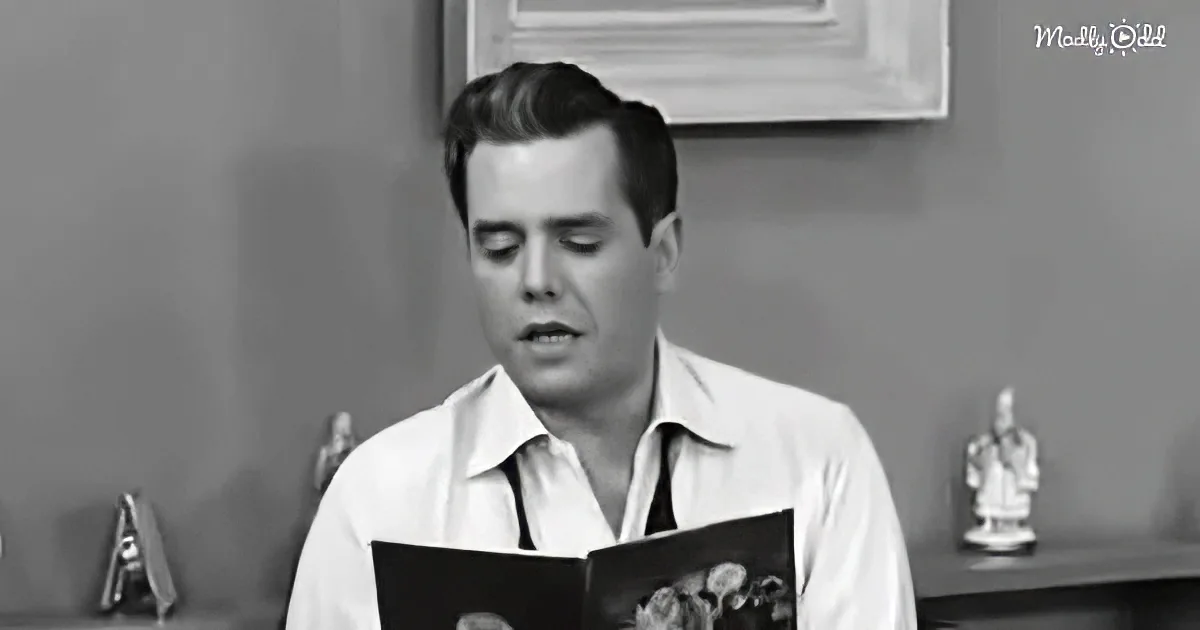 A man in a white shirt and tie reads a book, with a framed picture and small statue in the background.