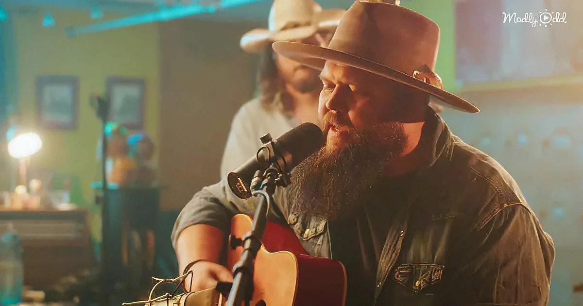 A man with a beard and cowboy hat sings into a microphone while playing guitar.