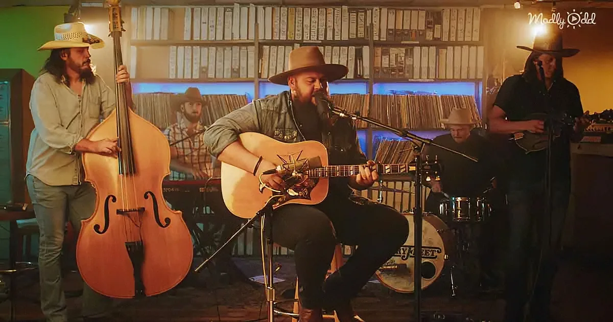 A man in a hat plays guitar and sings into a microphone in a band setting.
