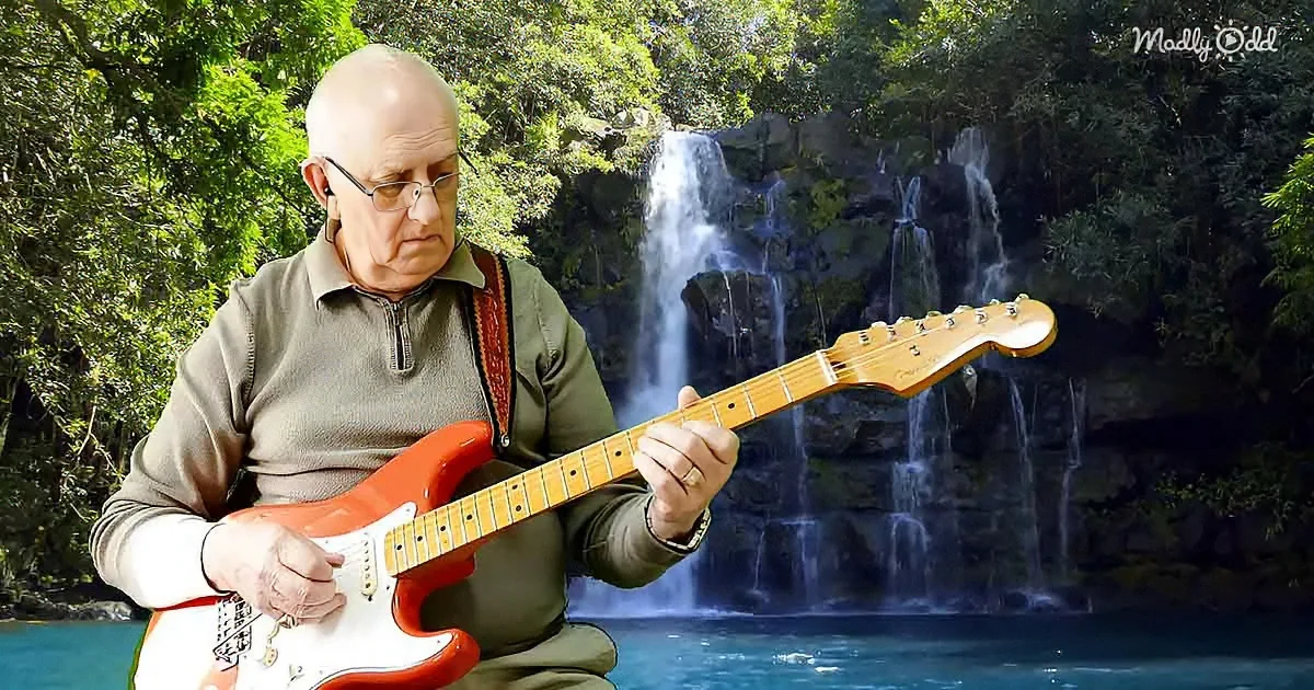 An elderly man plays a red electric guitar in front of a serene waterfall surrounded by lush greenery.