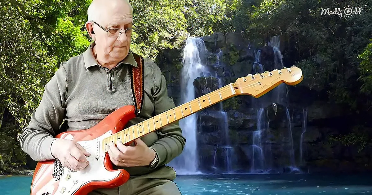 An older man plays a red electric guitar in front of a lush waterfall.