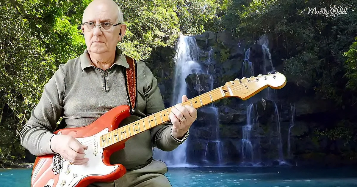 An older man plays a red electric guitar in front of a waterfall surrounded by lush greenery.