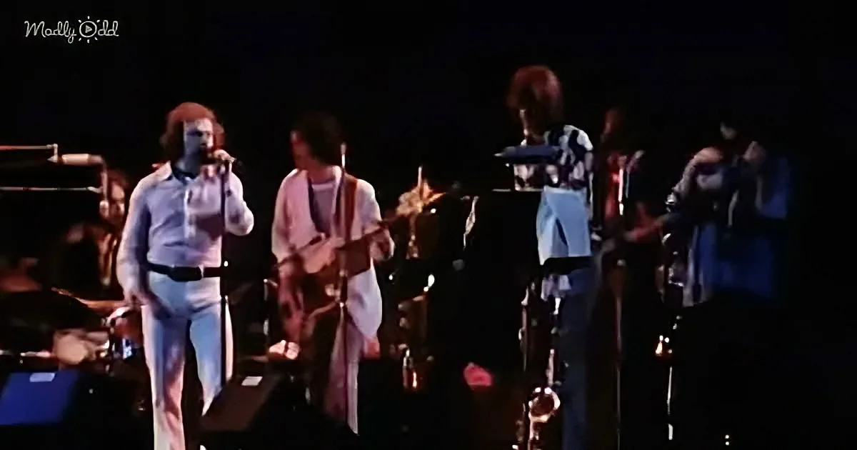A man in white plays guitar on stage with a band, microphone in front of him.