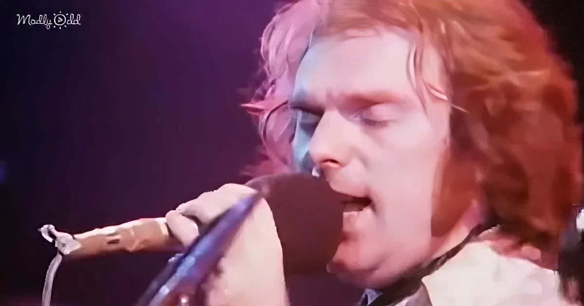 A person with red hair sings passionately into a microphone, illuminated by stage lights.