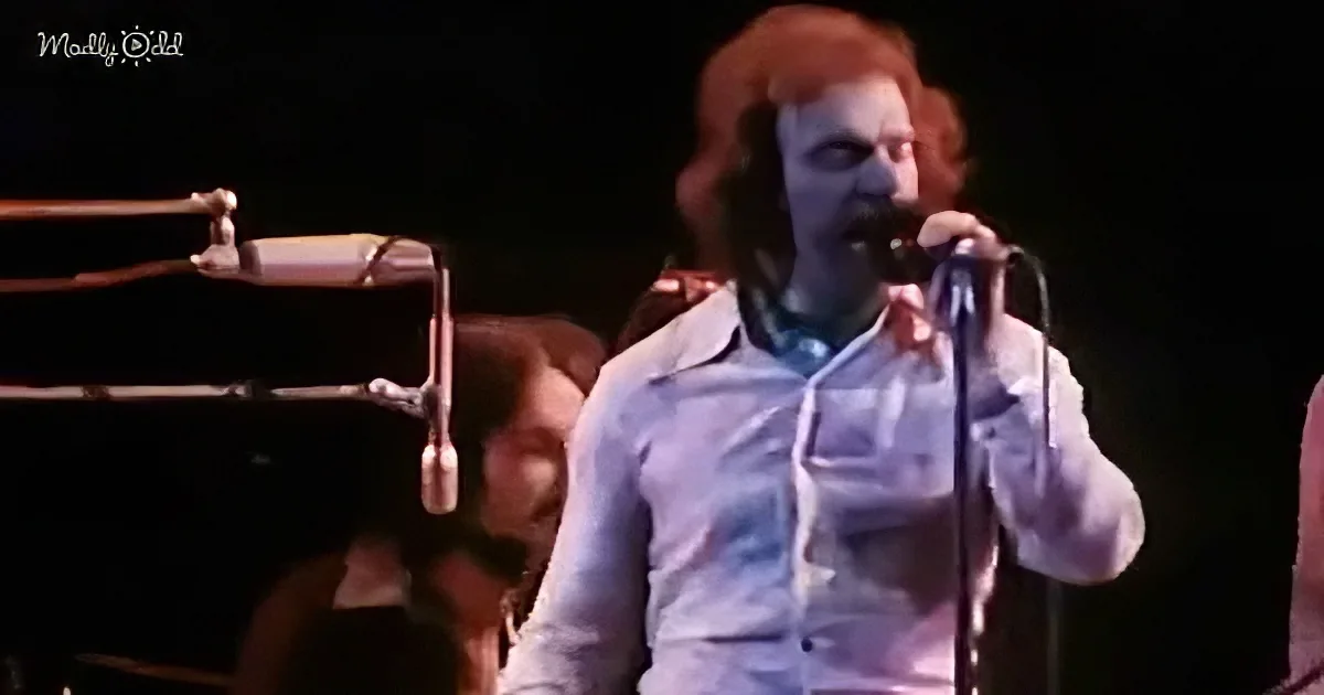 A person in a white shirt sings into a microphone on stage, with a keyboard and another person in the background.