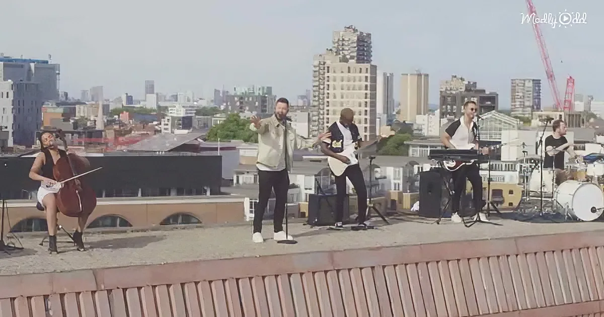 A band performs on a rooftop with a cityscape backdrop, featuring a cellist, guitarist, keyboardist, and drummer.