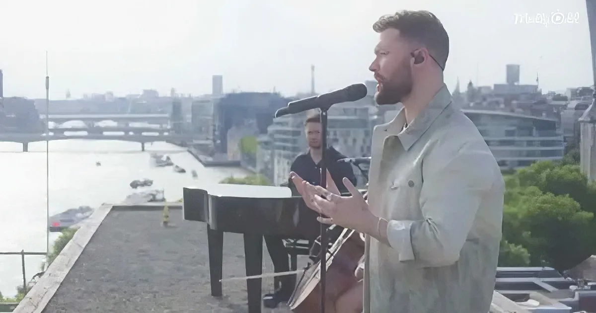 A man sings into a microphone on a rooftop with a cityscape and river in the background.