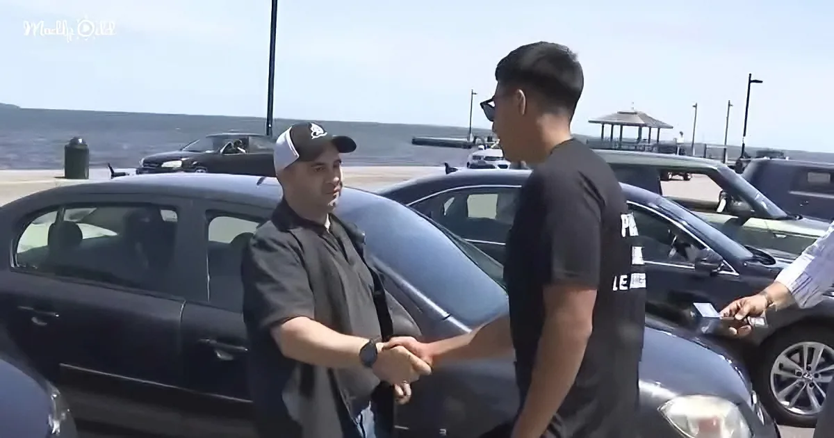 Two men shake hands in a parking lot near parked cars, with a scenic view in the background.