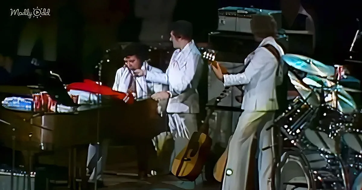 Musicians in white suits perform on stage with guitars and drums.