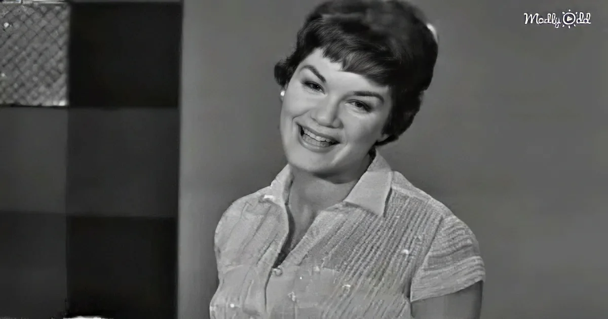 A smiling woman in a striped shirt, likely from a vintage TV show, with a blurred background.