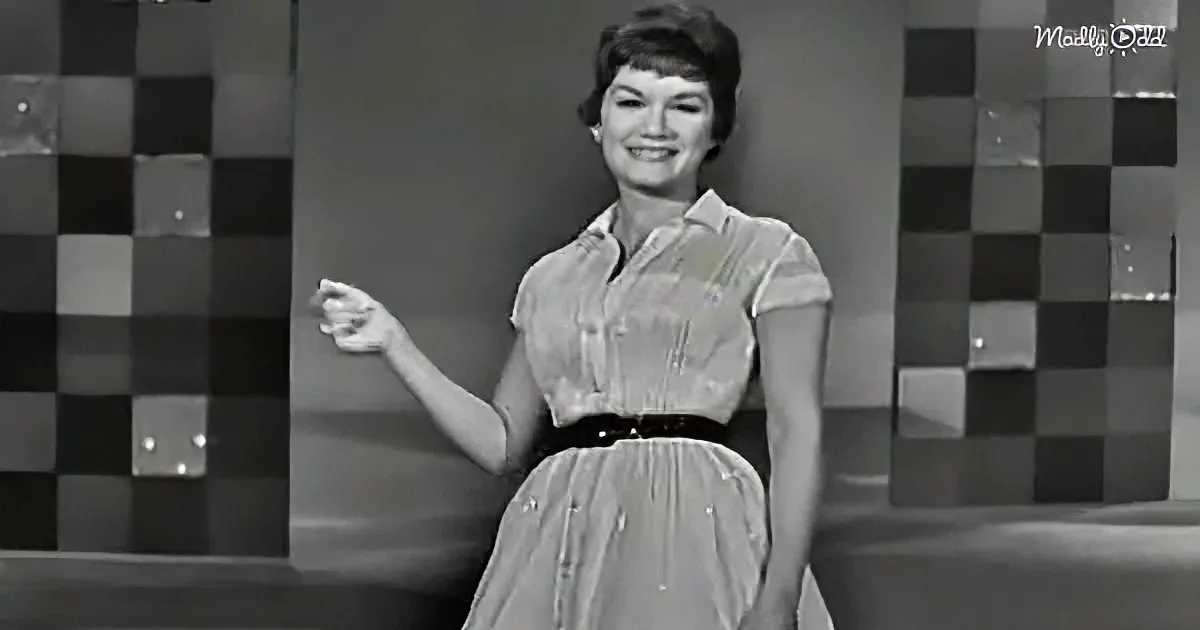 A woman in a 1950s-style dress smiles, gesturing with her right hand, against a checkered backdrop.