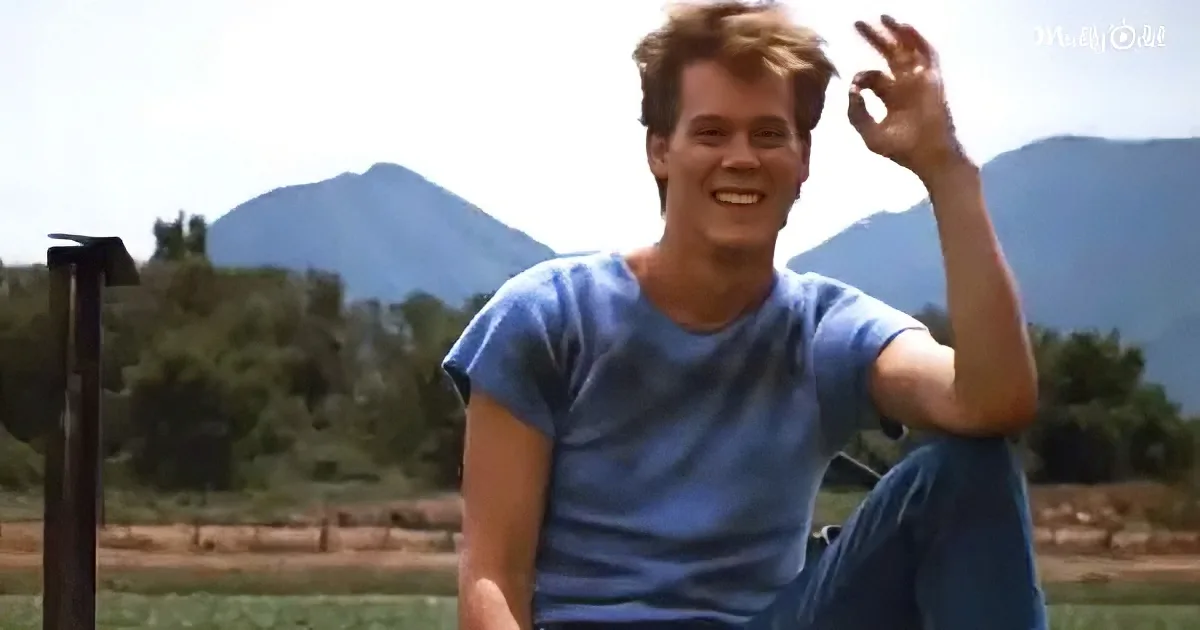 A man in a blue shirt makes an "OK" sign with his hand, smiling, with mountains and trees in the background.