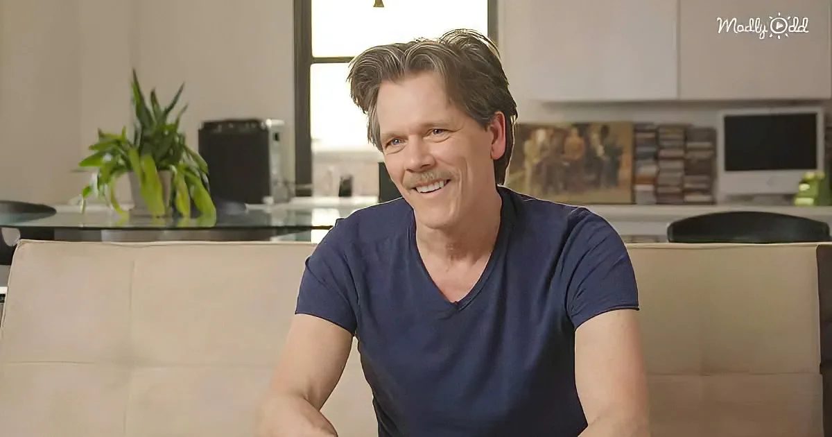 A man in a blue t-shirt sits on a beige couch, smiling, with a kitchen and plant in the background.