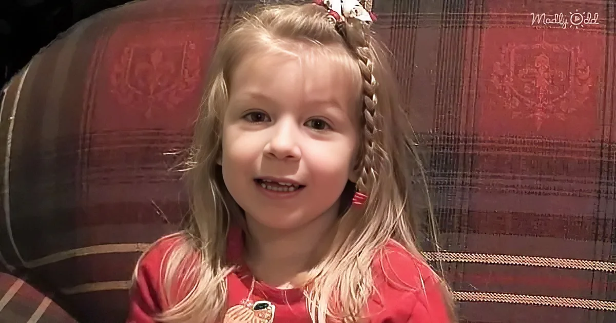 A young girl with long blonde hair in braids, wearing a red shirt, sits in front of a red plaid background.