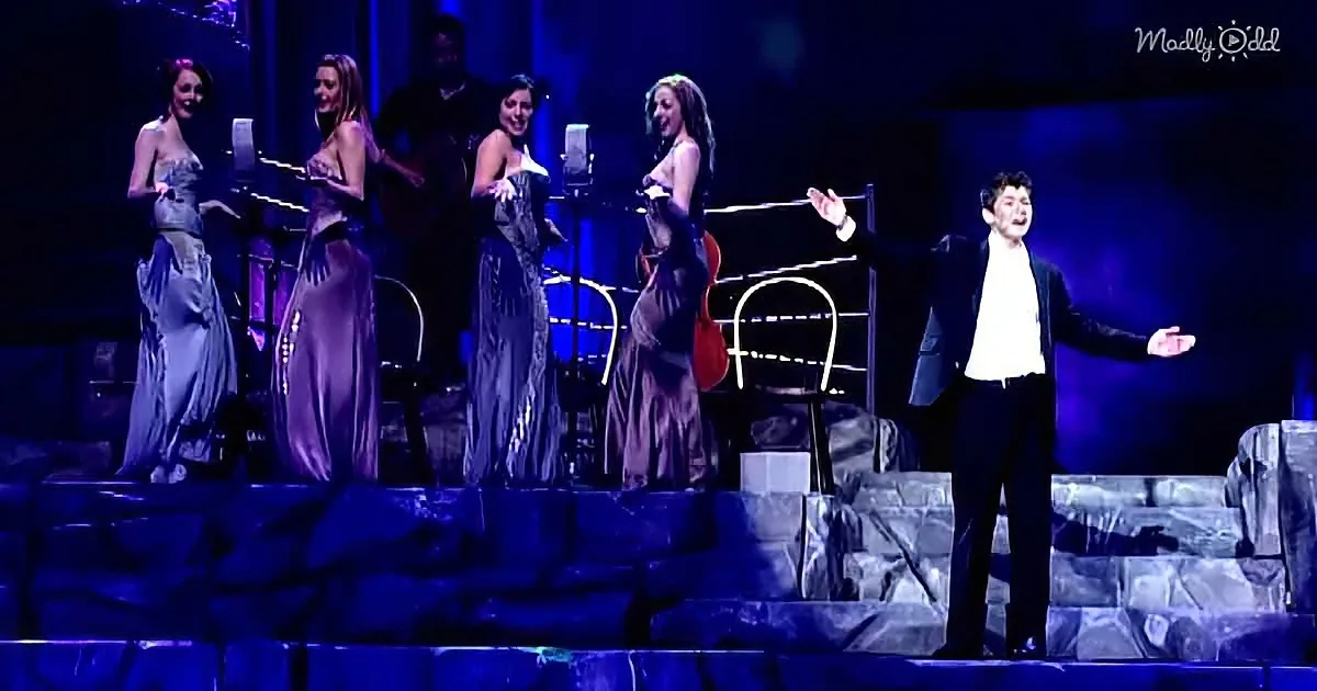 A man in a suit sings passionately on stage with four women in elegant dresses, accompanied by a guitarist.