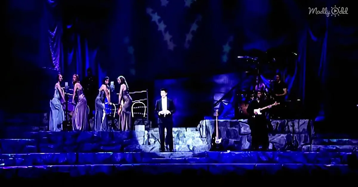 A man in a suit stands center stage, surrounded by women in elegant dresses and musicians, under blue lighting.