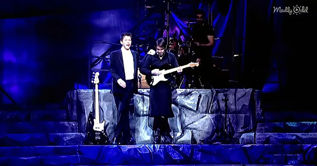 Two musicians perform on stage, one singing, the other playing guitar, under blue lighting.
