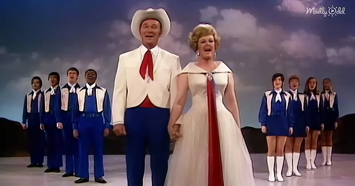 A man in a cowboy hat and white suit stands with a woman in a white dress and red sash, surrounded by a choir in blue...