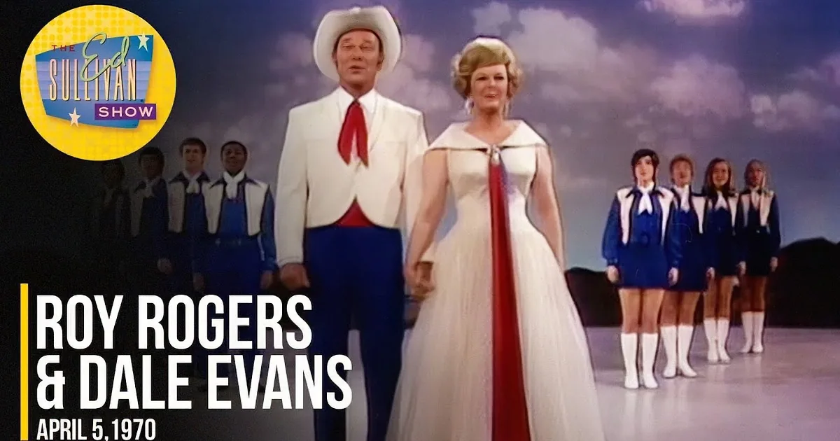 A man in a white suit and cowboy hat smiles alongside a woman in a white dress with blonde hair styled in a 1960s fashion.