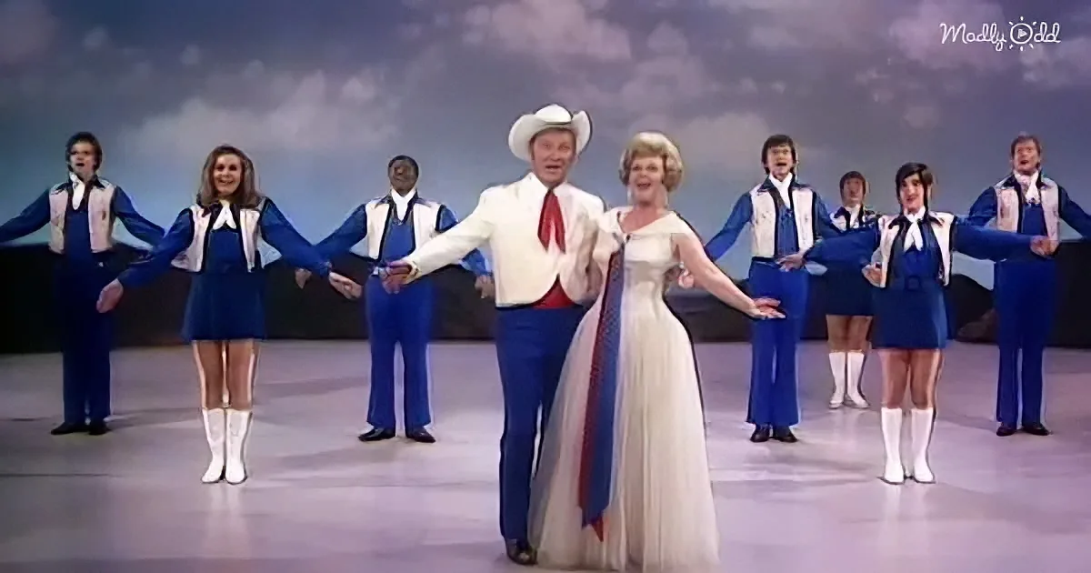 A man in a white suit and cowboy hat dances with a woman in a white dress, surrounded by a group in blue and white outfits.