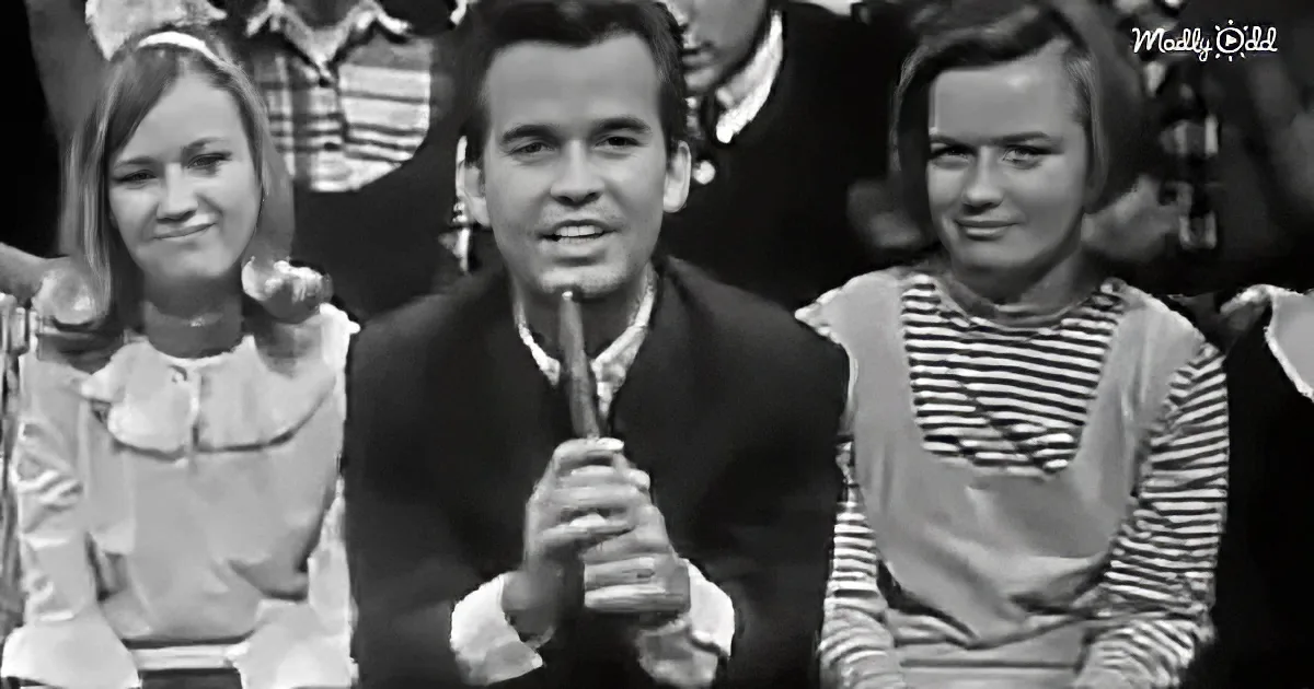 A man in a suit holds a microphone, flanked by two women in striped shirts, in a vintage setting.